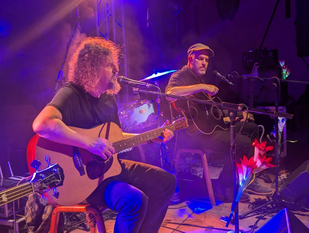 Andre Dusk an der Gitarre sowie der zweite Gitarrist der Band im Hintergrund auf der Bühne, die in blau-lilanes Licht getaucht ist.